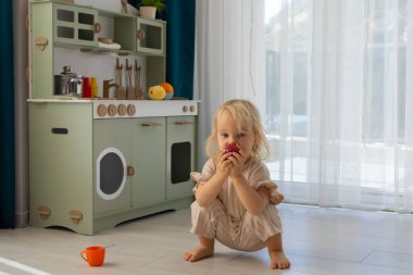 Yeni yürümeye başlayan bir çocuk, canlı bir oyun odasında elmanın tadını çıkarırken yerde rahatça oturuyor. Güneş ışığı perdelerin arasından akar ve yakınlardaki neşeli bir mutfak düzenini aydınlatır..