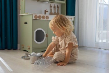 Pijama pantolonlu bir çocuk, kapalı bir alanda açık ahşap bir zeminde otururken beyaz yumurtalarla dolu bir tencereyle etkileşime geçmek için kaşık kullanır. Arka planda çocuk mutfağı. Rol yapma oyunları