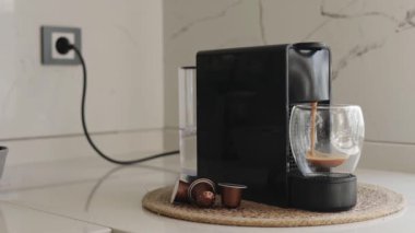 Şık, modern bir mutfakta, kapsül makinesi zengin bir espresso yapıp cam bardağa doldurur. Kurulumda, sabah evde ideal bir kahve anı gösteren kapsüller ve su deposu yer alıyor.