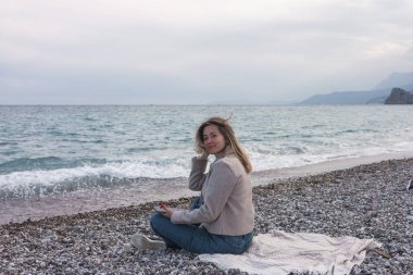 Yan bakış genç, gülümseyen kadın bej ceket ve kot pantolon giyiyor. Günlük giysiler giyiyor. Cep telefonu kullanıyor. 