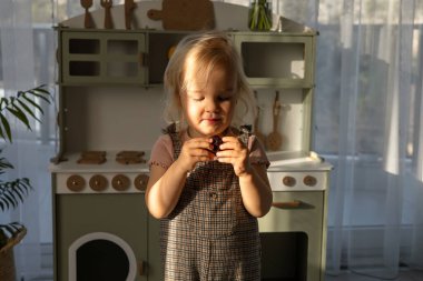 Neşeli genç bir kız ahşap mutfak setinin önünde elinde oyuncak yiyecekle duruyor. Çocuk gelişimi ve öğrenimi için çevre dostu ve güvenli el yapımı ahşap ürünler. 