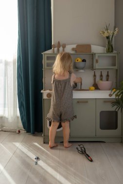 Genç bir kız yerde oturuyor, gülümsüyor ve oyuncaklarla dolu ahşap bir oyun mutfağını keşfediyor, pencereden süzülerek öğleden sonra ışığının tadını çıkarıyor..