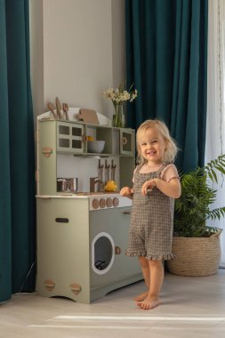 Kareli elbiseli küçük bir çocuk oyuncak mutfağı keşfediyor, gülümsüyor ve ahşap kap kacak ile meşgul oluyor. Çocuk gelişimi ve öğrenimi için çevre dostu ve güvenli el yapımı ahşap ürünler. 