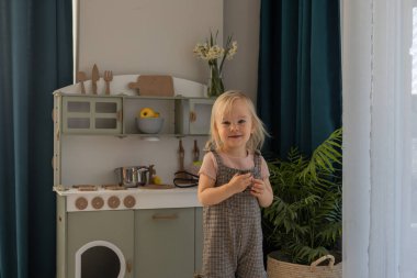 Çocuk, dekore edilmiş, aydınlık bir odada mutfak oyuncaklarıyla oynarken gülümsüyor. Minyatür mutfak çeşitli mutfak aletleri ve renkli eşyalar içeriyor. Çevre dostu ve güvenli el yapımı.