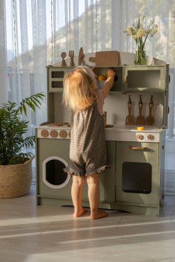 Küçük bir çocuk, aydınlık ve havadar bir odada yemek pişiriyormuş gibi yaparak küçük bir mutfak setinde duruyor. Çocuk gelişimi ve öğrenimi için çevre dostu ve güvenli el yapımı ahşap ürünler. 