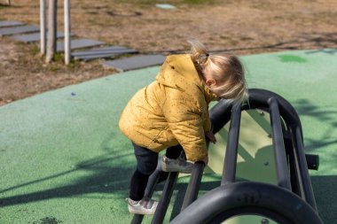 Küçük çocuk, sonbaharda bir parkta enerji dolu bir şekilde oyun parkına tırmanıyor. Manzaranın arka planında renkli ağaçlar ve görkemli dağlar bulunur. Egzersiz ekipmanlı kamusal alan