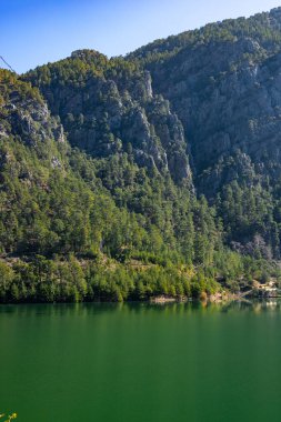 Sakin bir dağ gölü çevreleyen ağaçların ve sarp kayalıkların canlı yeşil yapraklarını yansıtır ve sabah ışığında açık mavi bir gökyüzünün altında doğanın güzelliğini gösterir. Türkiye, Antalya