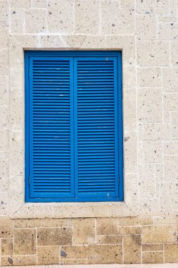 Mavi kepenkler açık renkli taştan bir duvarı tamamlıyor, sakin bir kıyı ortamında mimari detaylar sergiliyor. Fener, sakin atmosfere cazibe katıyor. Geleneksel Yunan renkleri ve stye