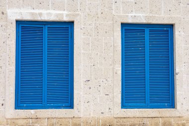 Mavi kepenkler açık renkli taştan bir duvarı tamamlıyor, sakin bir kıyı ortamında mimari detaylar sergiliyor. Fener, sakin atmosfere cazibe katıyor. Geleneksel Yunan renkleri ve stye