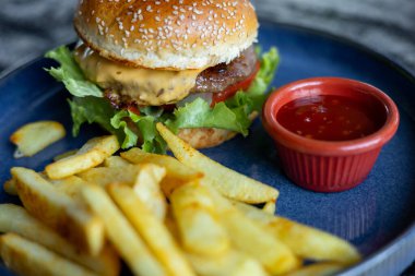 Lezzetli bir çizburger, üzerinde taze marul ve bir dilim peynirle mavi bir tabakta oturur. Yanında küçük bir kase tatlı sosu da eklenince yanında altın patates kızartması oluyor..