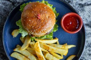 Marul ve kirazla kaplı lezzetli bir hamburgerin en üst görüntüsü altın patates kızartması ve küçük bir kase sosun yanında duruyor. Canlı renkler koyu plaka ve desenli yüzey ile tezat oluşturuyor. Fast food