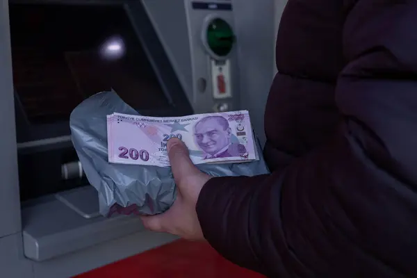 Türkiye, Antalya - 21.02.2025: Adam ATM 'den para çekiyor. Koruyucu bir çarşafa sarılı Türk lira banknotları tutuyor. Makine parlak kırmızı bir dış görünüş sergiliyor