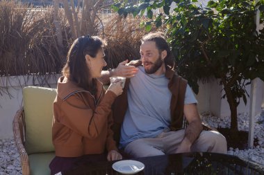 Bahçedeki pelüş bir kanepede oturan iki kişi sıcak bir sohbete dalıp kahvelerini yudumluyorlar. Güneş ışığı yeşil yaprakların arasından süzülür, dingin bir atmosfer yaratır, rahatlamak için mükemmeldir..