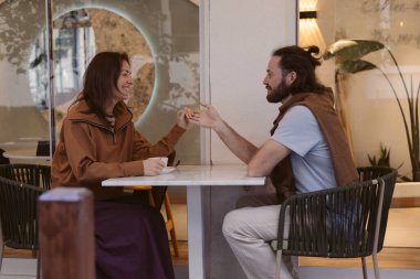 Bir çift şık bir kafede karşılıklı oturuyor, gülümsüyor ve sıcak bir sohbete girişiyor. Atmosfer, kapalı yeşil alan ve yumuşak ışıklandırma ile rahatladı. Ellerini tutuyorlar.