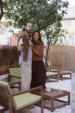 Erkek ve kadın açık havada bir ağacın altında samimi bir sohbet ederler. Dekor modern oturma, toprak renkleri ve rahatlamış bir atmosfer içeriyor, bağlantı için mükemmel. Kafede tarih