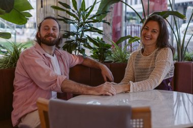 Genç bir aile büyük pencereli ve yemyeşil bitkileri olan şık bir kafede canlı bir sohbet ediyor. Atmosfer sıcak ve davetkar, rahat bir şehir ortamını yansıtıyor. Sevgililer birlikte vakit geçirir.