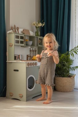 Kareli elbiseli küçük bir çocuk oyuncak mutfağı keşfediyor, gülümsüyor ve ahşap kap kacak ile meşgul oluyor. Çocuk gelişimi ve öğrenimi için çevre dostu ve güvenli el yapımı ahşap ürünler. 