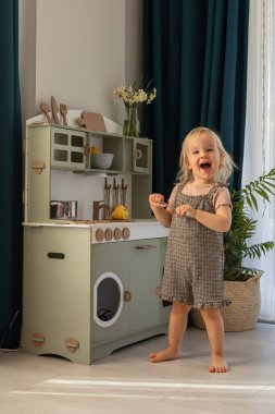 Kareli elbiseli küçük bir çocuk oyuncak mutfağı keşfediyor, gülümsüyor ve ahşap kap kacak ile meşgul oluyor. Çocuk gelişimi ve öğrenimi için çevre dostu ve güvenli el yapımı ahşap ürünler. 
