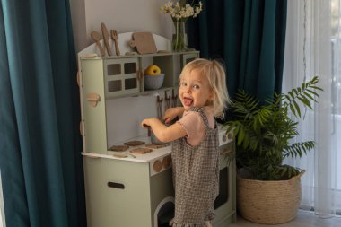 Genç çocuk renkli bir mutfak setinde bir tencereyi zevkle karıştırır. Güneş ışığı pencereden süzülüyor. Çocuk gelişimi ve öğrenimi için çevre dostu ve güvenli el yapımı ahşap ürünler. 