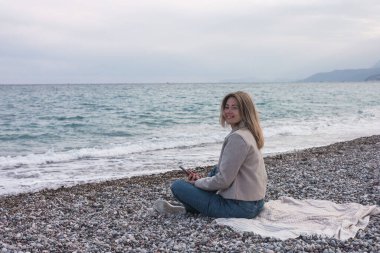 Yan bakış genç, gülümseyen kadın bej ceket ve kot pantolon giyiyor. Günlük giysiler giyiyor. Cep telefonu kullanıyor. 