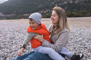 Bir anne ve küçük oğlu kışın çakıllı plajda oturuyorlar. Gülümsüyor ve birlikte oynuyorlar. Etrafı dağlarla çevrili ve sakin bir deniz..
