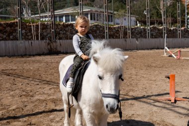 Genç bir kız, kumlu bir arenada pofuduk beyaz bir midillinin üzerinde güvenle oturuyor. Aydınlık bir gün ve çevre alan yeşil ve kırsal yapılarla kaplı. Eko at çiftliğinde aile günü