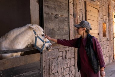 Bir insan elini nazikçe kırsal bir ahırın camına kafasını sokan beyaz bir midilliye yiyecek uzatır. Güneş ışığı sahneyi aydınlatır ve sıcak bir atmosfer yaratır..