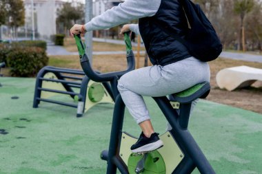Bir insan, halka açık bir parkta egzersiz aletleri üzerinde çalışıyor. Kadınlar günlük spor kıyafetleri giyer ve sakin bir ortamda antrenmanlarına odaklanırlar..