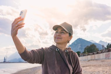 Şapka takan bir kadın dışarıda duruyor, akıllı telefonuyla özçekim yapıyor. Arka planda görkemli dağlar ve bulutlu bir gökyüzü var. Güzel bir atmosfer yaratıyorlar.. 