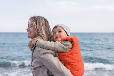 Kışın kumsalda bir anne ve oğlu birlikte güzel vakit geçiriyorlar. Kıyı şeridi boyunca çakıl taşları toplarken çocuk annelerinin sırtında mutlu bir şekilde at sürüyor. Mutlu ve eğlenceli aile zamanı.