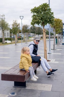 Bir anne ve küçük kızı halka açık bir parkta bir bankta oturuyorlar. Ağaçlar ve açık alan sakin bir atmosfer yaratırken onlar birlikte vakit geçiriyorlar, gülümsüyorlar ve etkileşime geçiyorlar..
