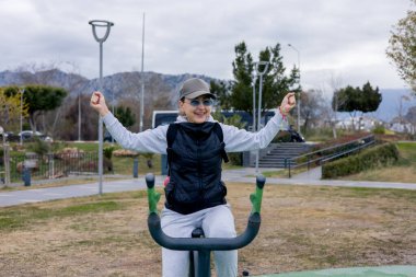 Son sınıf öğrencisi bir kadın parktaki açık hava spor aletleriyle antrenman yapıyor. Kendini yarışmanın galibi olarak hayal ederek ellerini kaldırdı. Etrafında ağaçlar ve bulutlu bir gökyüzü var..