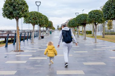 Ebeveyn büyükanne, ağaçlarla kaplı geniş bir gezinti alanında küçük bir çocukla el ele yürüyor. Çocuk, sarı ceketli, bulutlu bir günde sevdikleriyle anın tadını çıkartıyor..
