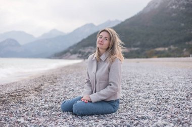 Sarışın kadın çakıllı sahilde oturuyor. Kış manzarası bulutlu bir gökyüzünün altında arka planda dağlar barındırır..
