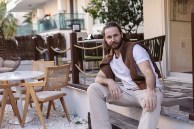 Uzun saçlı, bıyıklı ve sakallı yakışıklı adam merdivenlerde oturuyor, günlük kıyafetler giyiyor, arka planda oturma ve yeşillik olan modern bir açık hava ortamının tadını çıkarıyor..