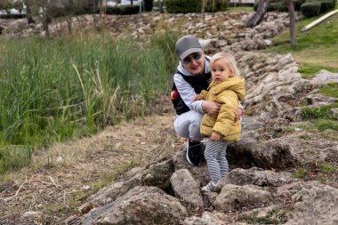 Bir büyükanne, torunuyla birlikte parkta su kenarında poz veriyor. Etrafları kayalar ve uzun otlarla çevrili. Açık havada sakin bir bahar gününün tadını çıkarıyorlar..