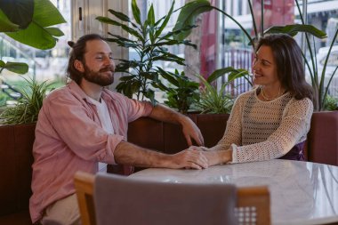 İki insan bir kafede, bitkilerle süslenmiş bir masada samimi bir anı paylaşıyor. El ele tutuşuyorlar, birbirlerine gülümsüyorlar, sıcak ve davetkar bir atmosfer yaratıyorlar. Genç bir ailenin günlük hayatı