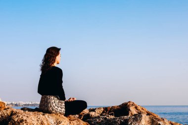 Genç bir kadın, sakin bir okyanusta güneş yükselirken huzur içinde kayalık bir sahilde meditasyon yapıyor. Açık gökyüzü ve sakin ortam yansıma için mükemmel bir atmosfer yaratır..