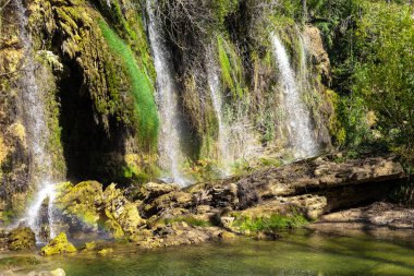 Çoklu şelaleler yosun kaplı kayaların üzerinden sakin bir havuza akıyor. Yeşillik bölgeyi çevreleyerek doğal ortamda barışçıl bir ortam yaratıyor..