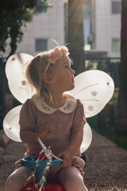 Çiçekli bir saç bandı ve kelebek kanatlı pembe bir elbise giyen küçük bir kız çocuğu güneşli bir bahçede oturmuş neşe saçıyor. Yumuşak ışık onun masumiyetini ve kaprisli kıyafetlerini vurguluyor..