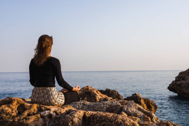 Dişi yogi, gün batımında okyanus ya da deniz kenarında yoga yapıyor. Genç kıvırcık kadının lotus yogası pozunda meditasyon yapması. Milenyum kızı kayalık sahilde yoga ve meditasyon yapıyor.