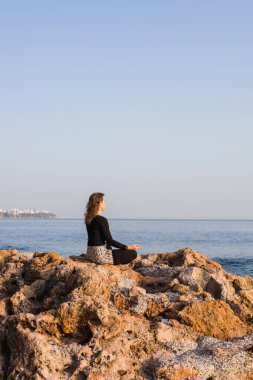 Genç bir kadın, kayalık sahilde ellerini bağlamış, nilüfer ağacında poz verirken meditasyon yapıyor. Deniz ya da okyanus yakınında bahar günbatımı. Rahatla ve huzur içinde yat.