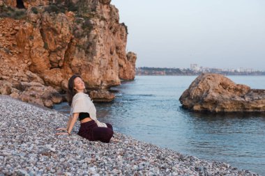 Genç bir kadın, kayalık sahilde ellerini bağlamış, nilüfer ağacında poz verirken meditasyon yapıyor. Deniz ya da okyanus yakınında bahar günbatımı. Rahatla ve huzur içinde yat.