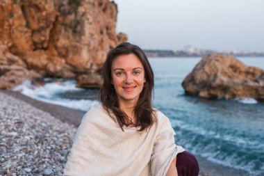 Bir kadın, kayalık kayalıklar ve nazik dalgalarla çevrili deniz kenarında huzurlu bir anın tadını çıkarır. Gün batımı, huzurlu manzaranın üzerine sıcak bir parıltı yayar ve rahatlatıcı bir atmosfer yaratır..