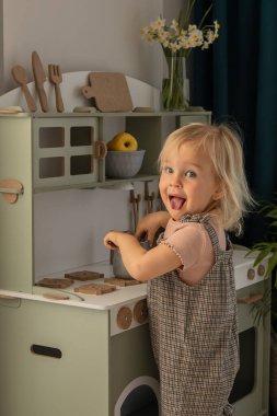 Genç çocuk renkli bir mutfak setinde bir tencereyi zevkle karıştırır. Güneş ışığı pencereden süzülüyor. Çocuk gelişimi ve öğrenimi için çevre dostu ve güvenli el yapımı ahşap ürünler. 