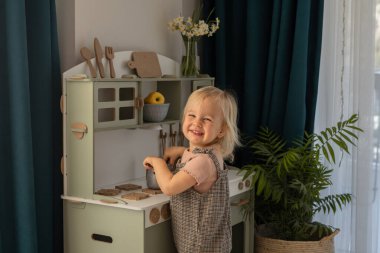 Genç bir çocuk, dekore edilmiş, aydınlık bir odada mutfak oyuncaklarıyla oynarken gülümsüyor. Minyatür mutfakta çeşitli mutfak aletleri ve renkli eşyalar var..