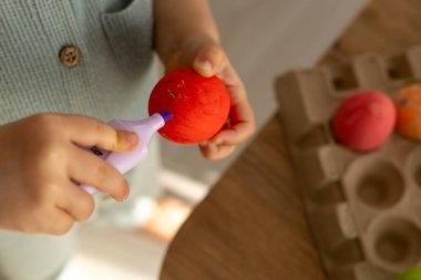 Bir çocuk dokuma sepetinden parlak renkli Paskalya yumurtalarını dikkatlice seçiyor. Sahne, bayram hazırlıklarına başlarken baharın neşeli ruhunu yakalıyor..