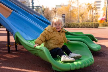 Sarı saçlı küçük bir çocuk yeşil bir kaydırağın üzerinde oturuyor ve aşağı kaymaya hazırlanırken gülümsüyor. Oyun alanı açık gökyüzünün altında sonbahar renkleriyle doludur..