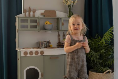 Çocuk, dekore edilmiş, aydınlık bir odada mutfak oyuncaklarıyla oynarken gülümsüyor. Minyatür mutfak çeşitli mutfak aletleri ve renkli eşyalar içeriyor. Çevre dostu ve güvenli el yapımı.