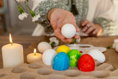 Bir insan, mum ve çiçeklerle çevriliyken Paskalya yumurtalarına renkli süsler çizer. Neşeli atmosfer bahar ruhunu ve bayram kutlamasını yansıtıyor..
